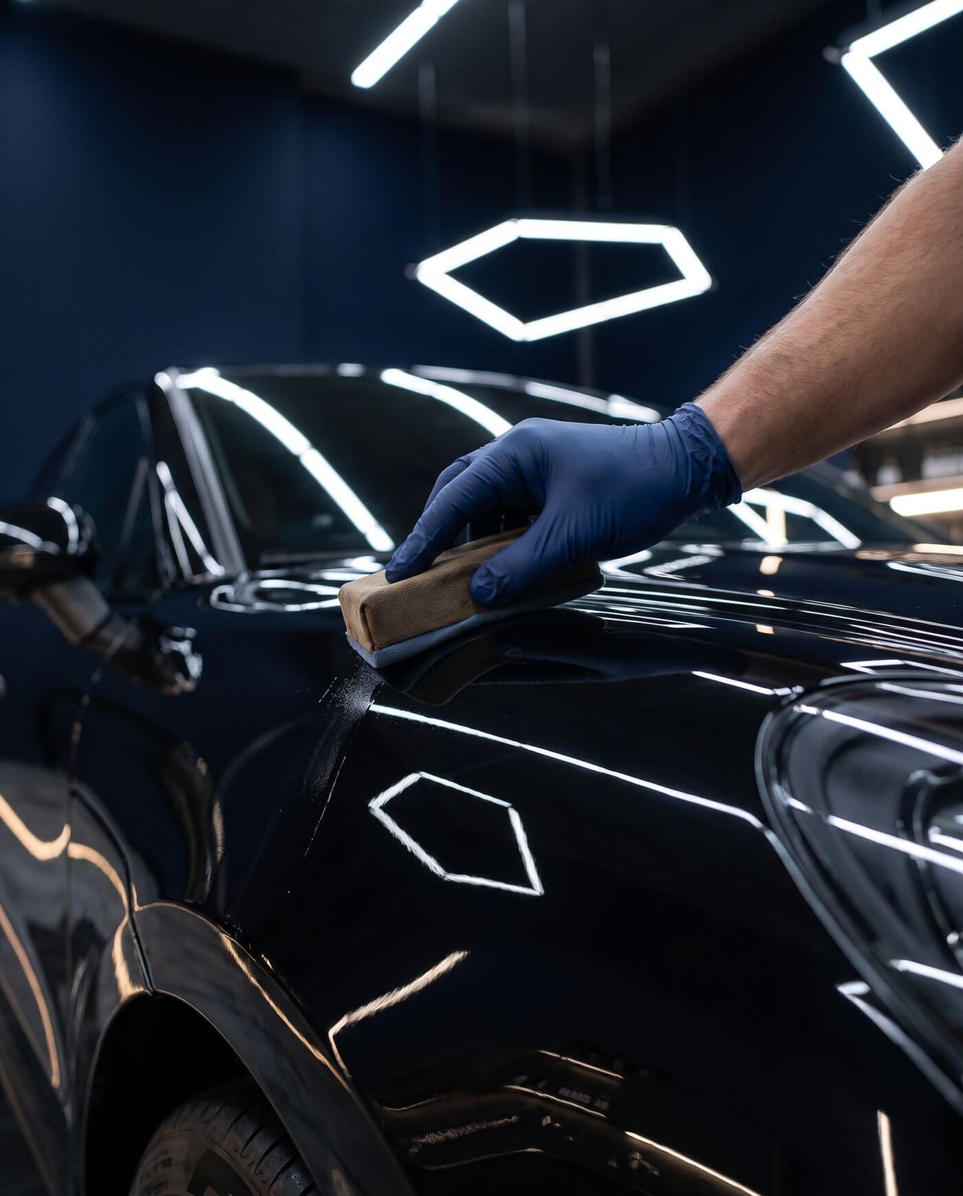 Mano con guante de nitrilo aplicando el revestimiento cerámico con una esponja sobre la carrocería negra de un coche, que refleja las luces del taller como un espejo